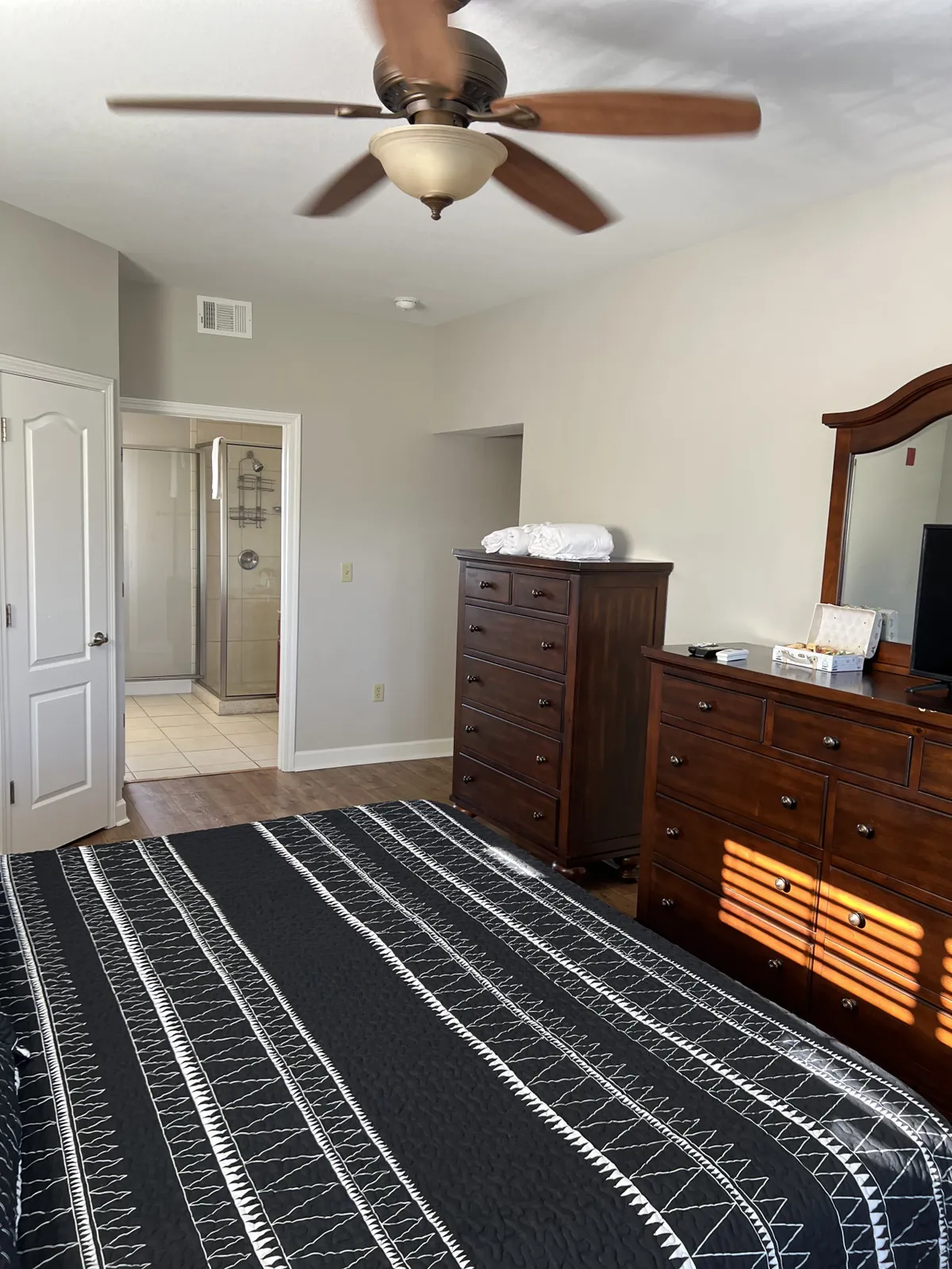 A serene bedroom with a bed featuring a patterned quilt, a classic wooden dresser, a ceiling fan overhead, and a glimpse of a shower area in the background. Soft natural light fills the room, creating a tranquil atmosphere ideal for relaxation.