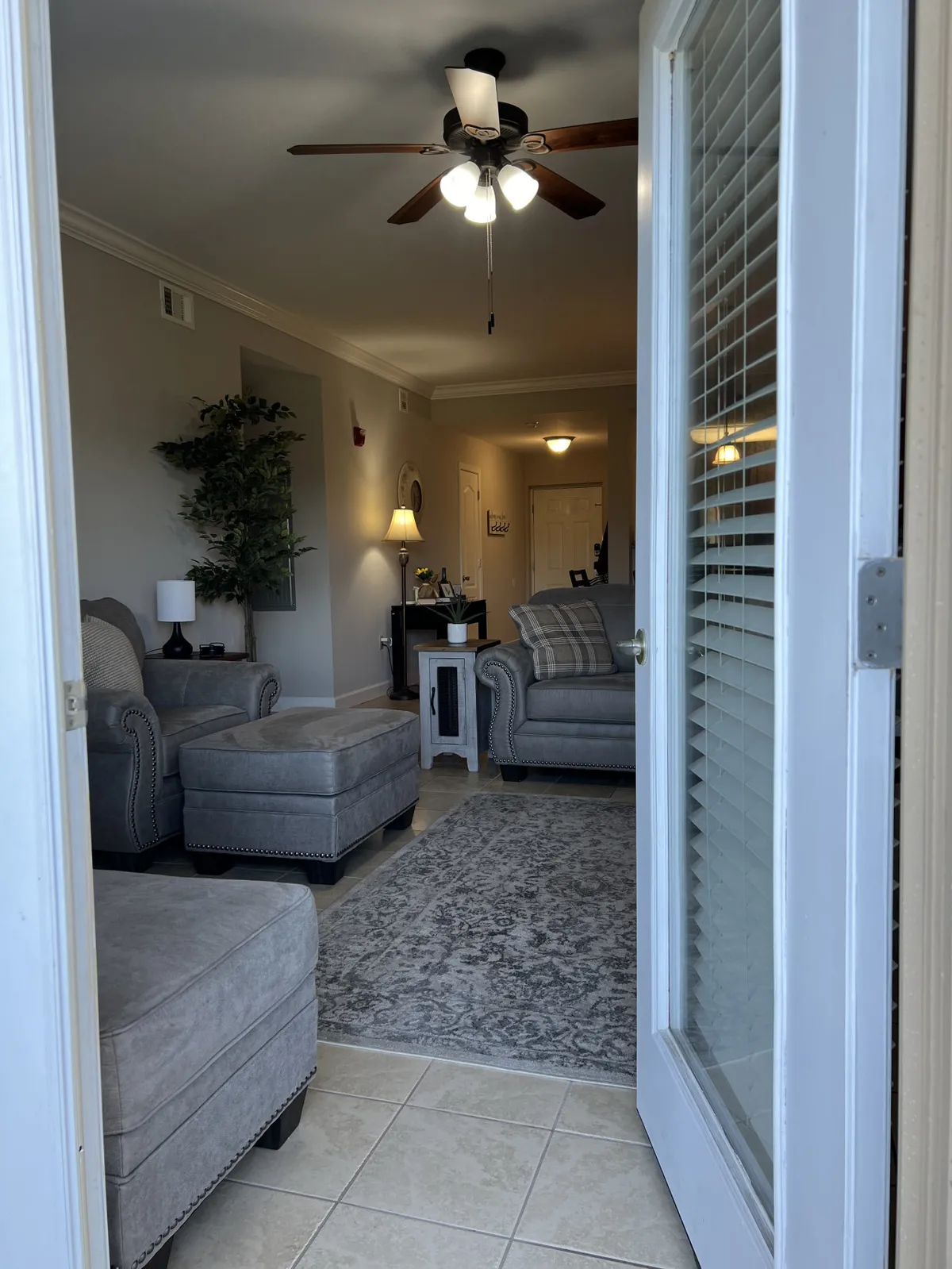 The photo shows the condo's entrance doorway leading into a comfortable living space with a sofa set, ceiling fan, decorative plant, and side table with a lamp. The space is bright, open, and ideal for welcoming family and friends.