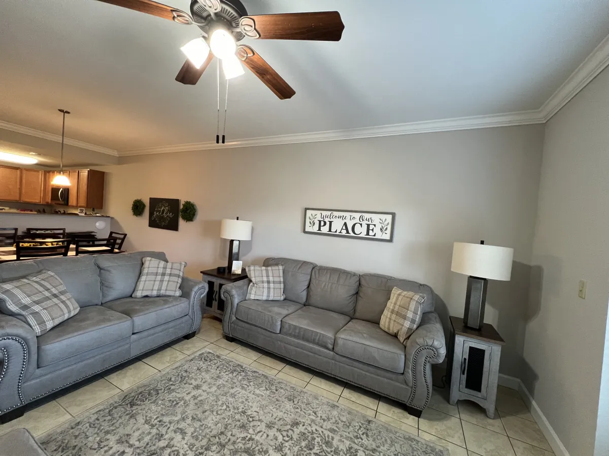 The scene features gray sofas with decorative pillows and a welcome sign, with a view of the dining area in the background. Table lamps and cozy lighting add to the inviting ambiance, suggesting a space designed for both relaxation and group gatherings.