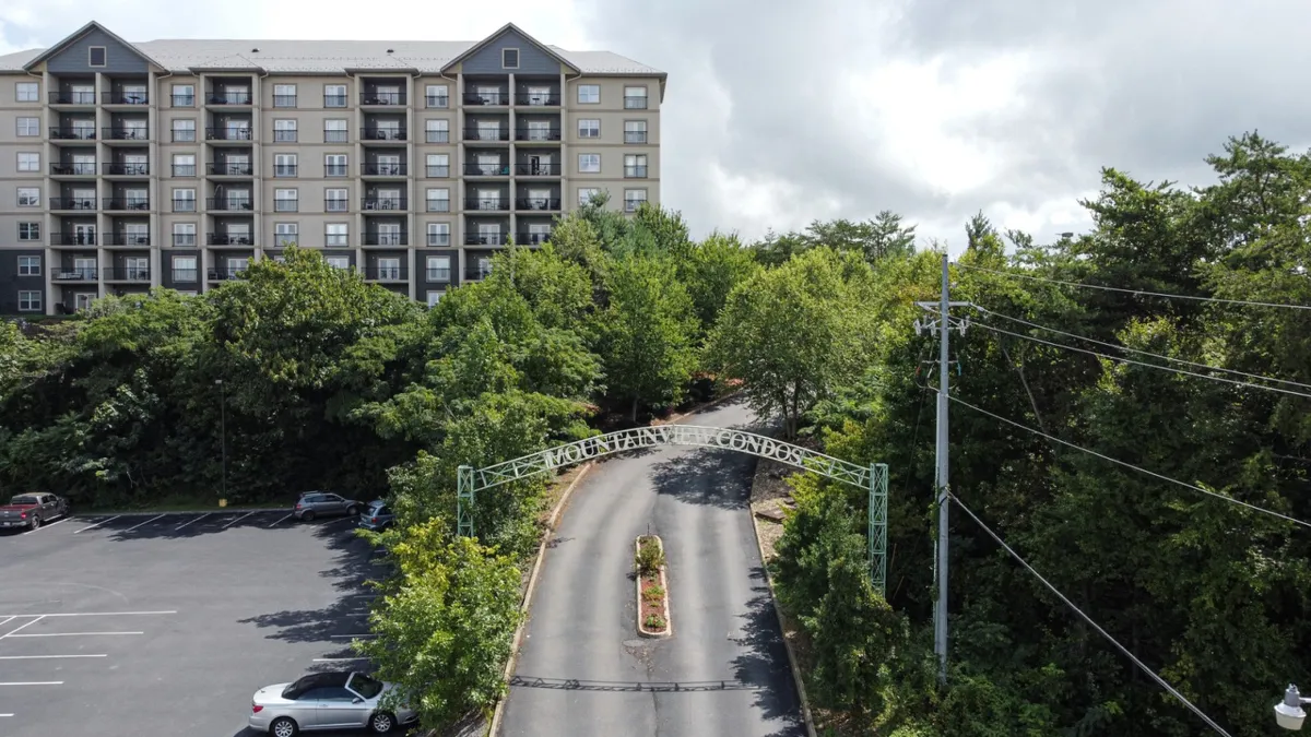 This image features the main entrance sign to the condominium community, a driveway leading to the condos, and lush surrounding greenery. The building is visible in the background, emphasizing a welcoming, resort-like atmosphere and a sense of arrival.