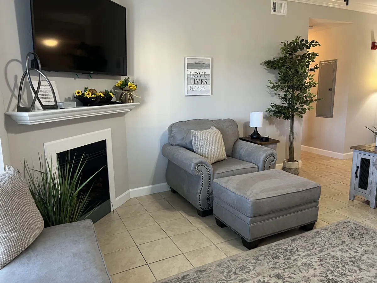 A welcoming living area featuring a comfortable armchair, decorative ottoman, fireplace, and lush indoor plant. The scene is set for relaxation or socializing, with ample natural light and inviting decor.